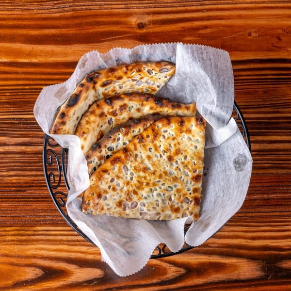 A basket of warm, charred flatbread with sesame seeds, lined with white paper, on a wooden table.