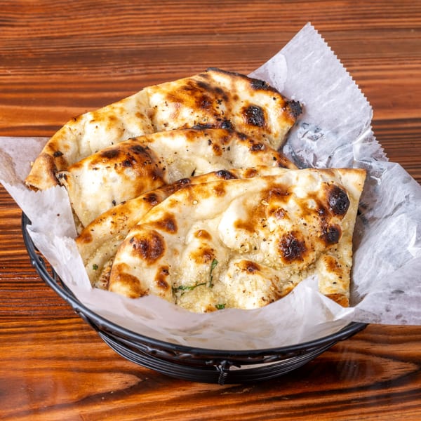 Golden brown naan bread, speckled with herbs and char marks, served in a black basket lined with parchment paper.