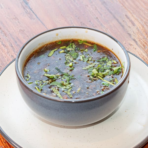 Dark brown soup with green herbs in a gray bowl on a white plate, on a wooden table.