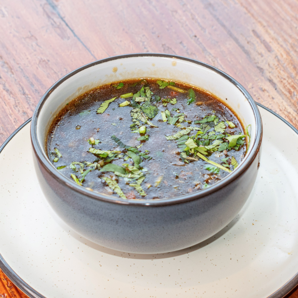 Dark brown soup with green herbs in a gray bowl on a white plate, on a wooden table.