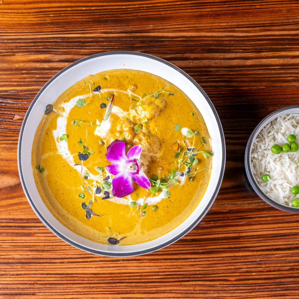 Golden curry with cream swirls, microgreens, and a purple orchid, served with a side of rice and peas.