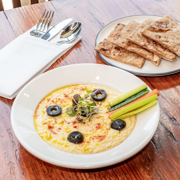 Hummus with olives, microgreens, and vegetable sticks, served with pita bread on a wooden table.