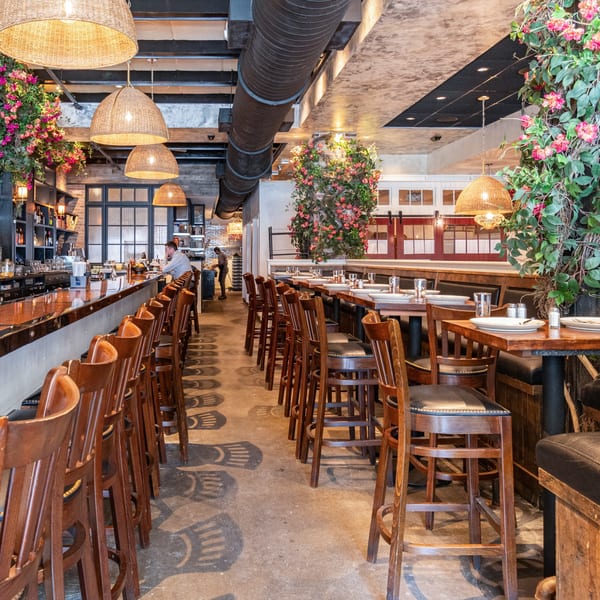 A restaurant interior with a long bar, high-top tables, and hanging floral decorations.