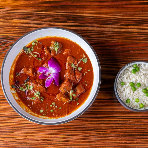 A bowl of red curry with meat, garnished with a purple orchid and microgreens, next to a bowl of rice with peas.