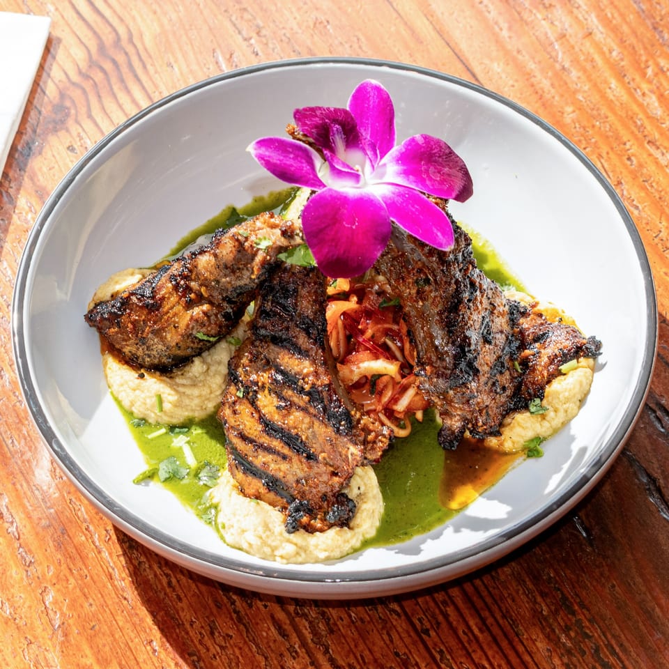 Grilled meat with a purple orchid garnish, served on a bed of hummus with green sauce.
