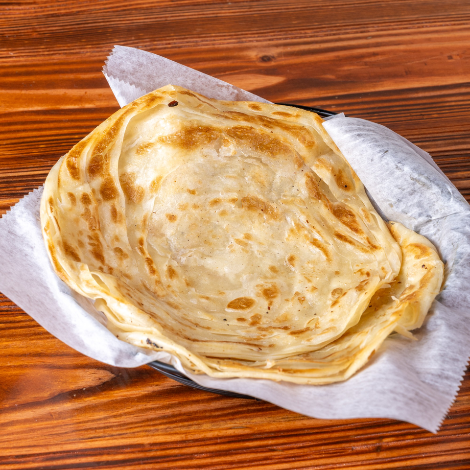 Golden brown paratha bread, folded and served in a basket lined with white paper on a wooden table.