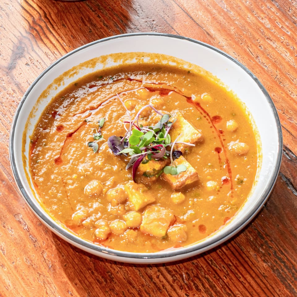 A bowl of chana masala with paneer and microgreens on a rustic wooden table.