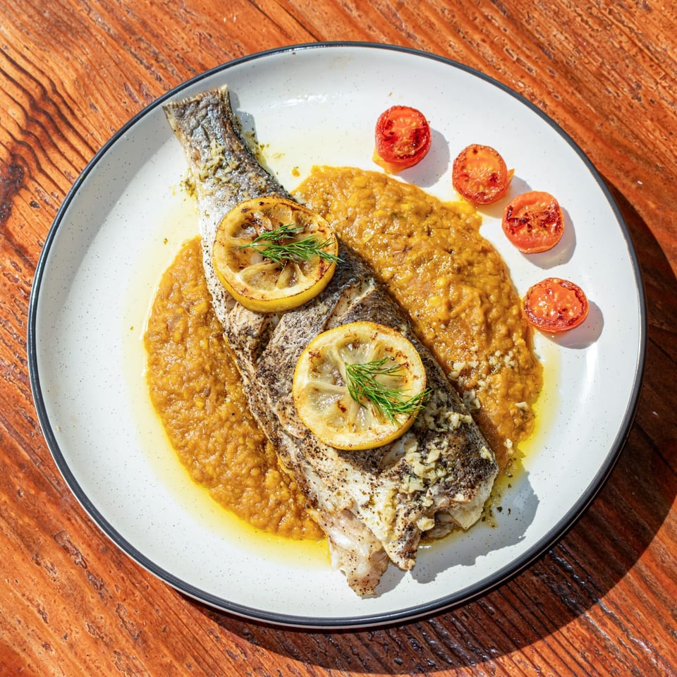 Whole roasted fish with lemon slices, dill, and roasted tomatoes on a bed of orange puree.