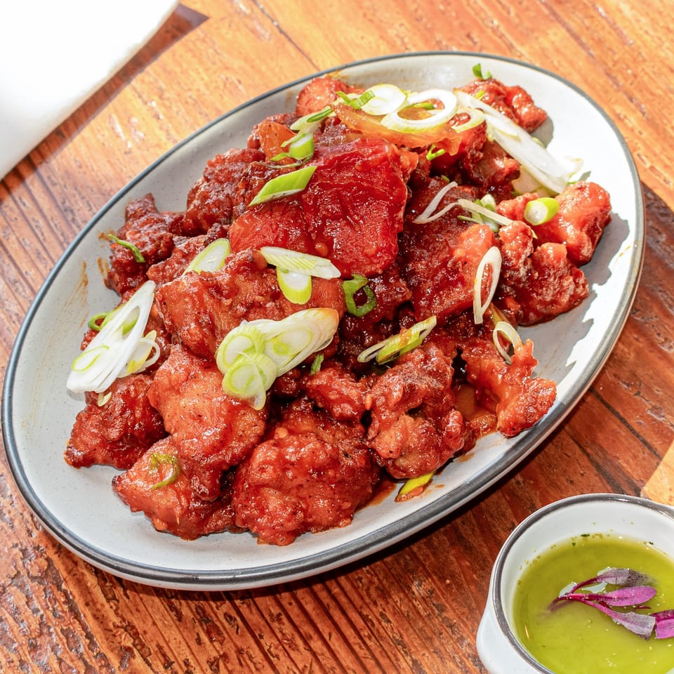 Sweet and sour chicken with sliced scallions on a white plate, next to a green dipping sauce.