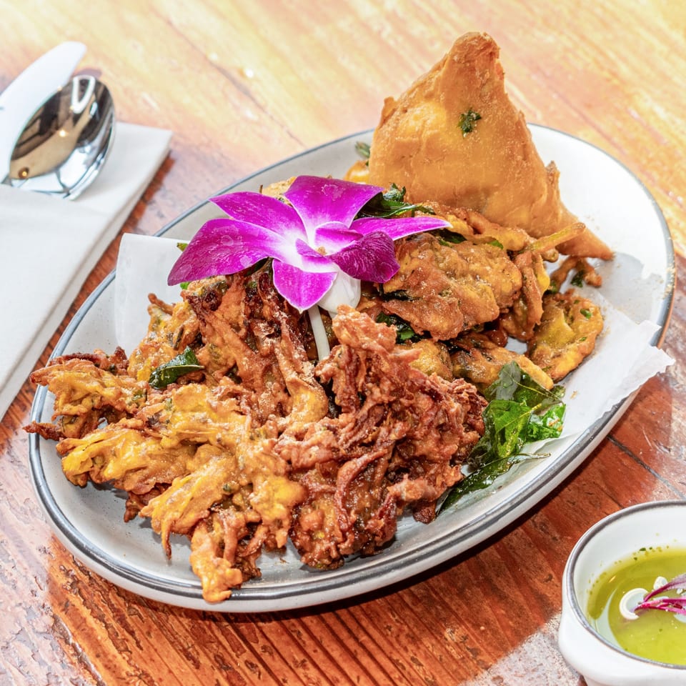 A plate of golden-brown pakoras and a samosa, garnished with a purple orchid, on a wooden table.