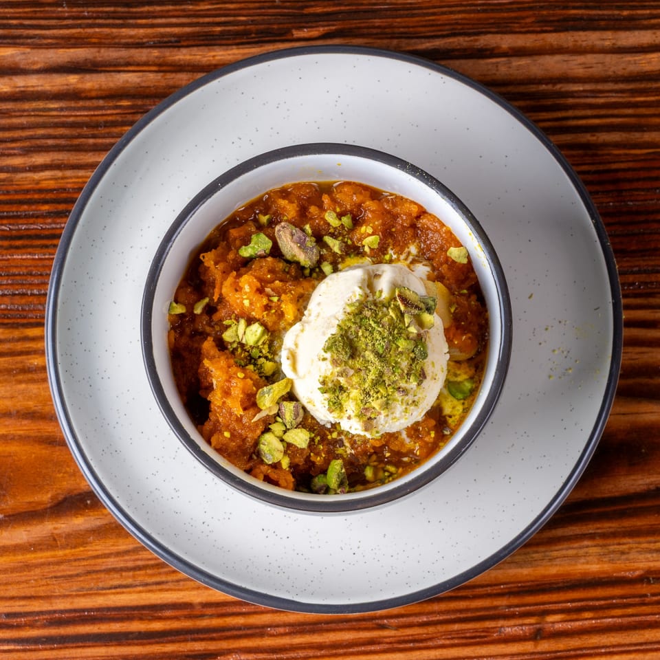 A bowl of orange halwa dessert topped with a scoop of vanilla ice cream and chopped pistachios.