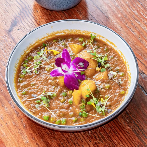 A bowl of curry with potatoes, peas, microgreens, and a purple orchid on a wooden table.