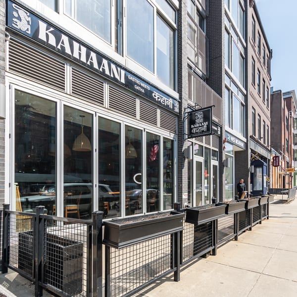 The exterior of Kahaani restaurant, featuring large windows, a black sign, and a fenced patio area.