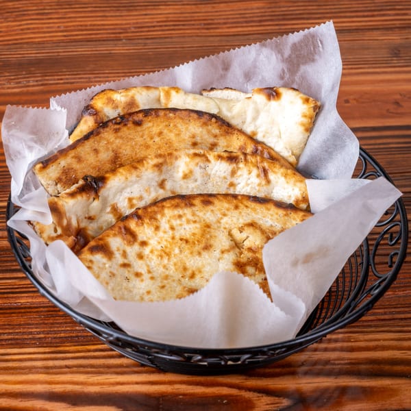 A black wire basket filled with several pieces of warm, golden-brown naan bread on a wooden table.