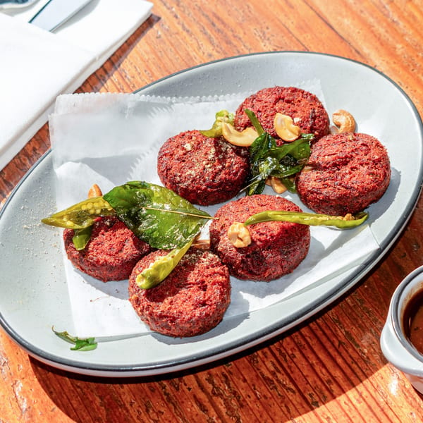 Six red beet cutlets garnished with cashews, green chilies, and curry leaves on a white plate.
