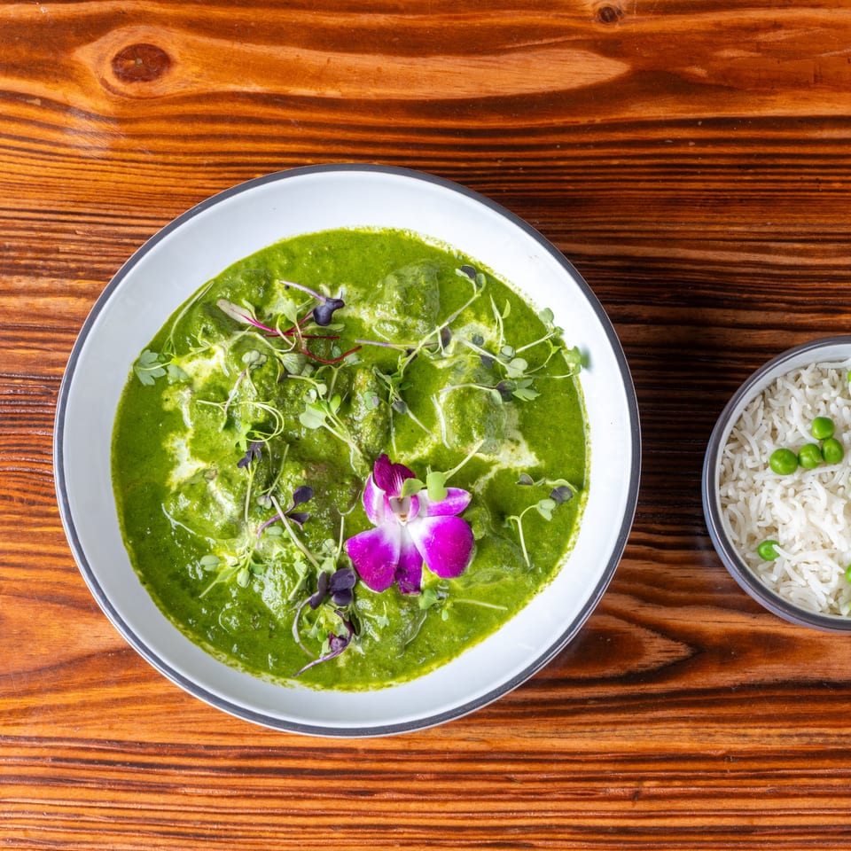 Palak Paneer, a vibrant green spinach curry with cheese, garnished with a purple orchid and microgreens, served with a side of rice and peas.