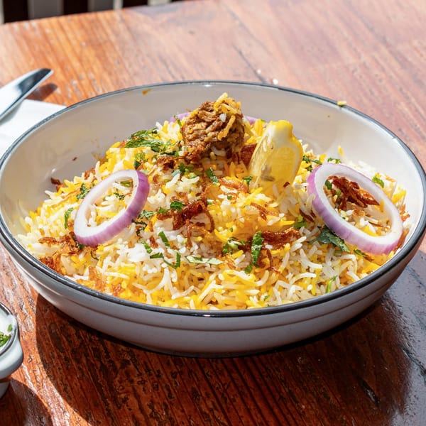 Close-up of a bowl of biryani with rice, meat, red onion, lemon, and herbs.