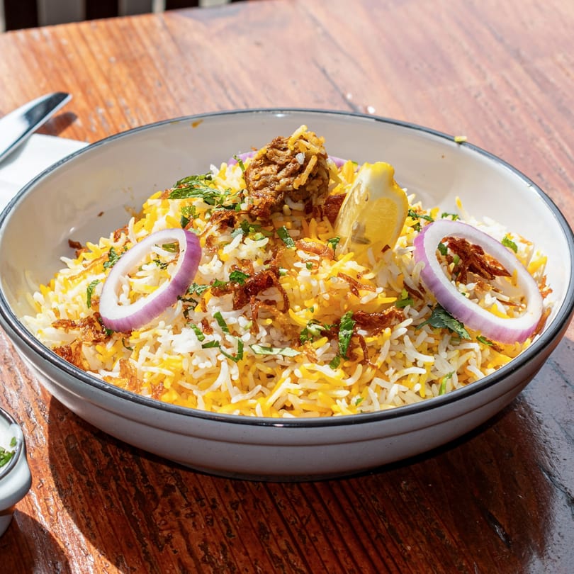 Close-up of a bowl of biryani with rice, meat, red onion, lemon, and herbs.