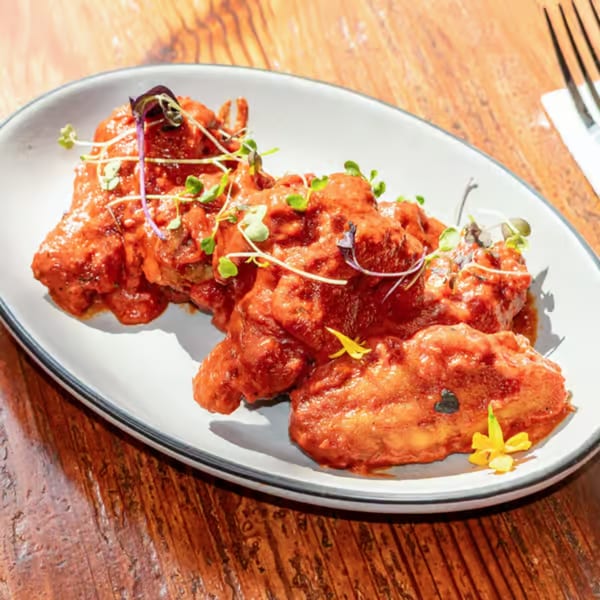 A white oval plate holds several chicken wings coated in a vibrant red sauce, garnished with microgreens.