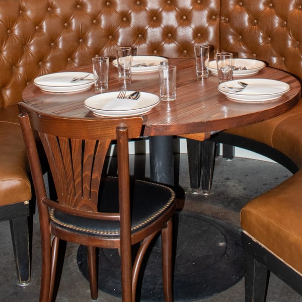 A round wooden table with place settings for six, surrounded by brown leather booth seating and a wooden chair.