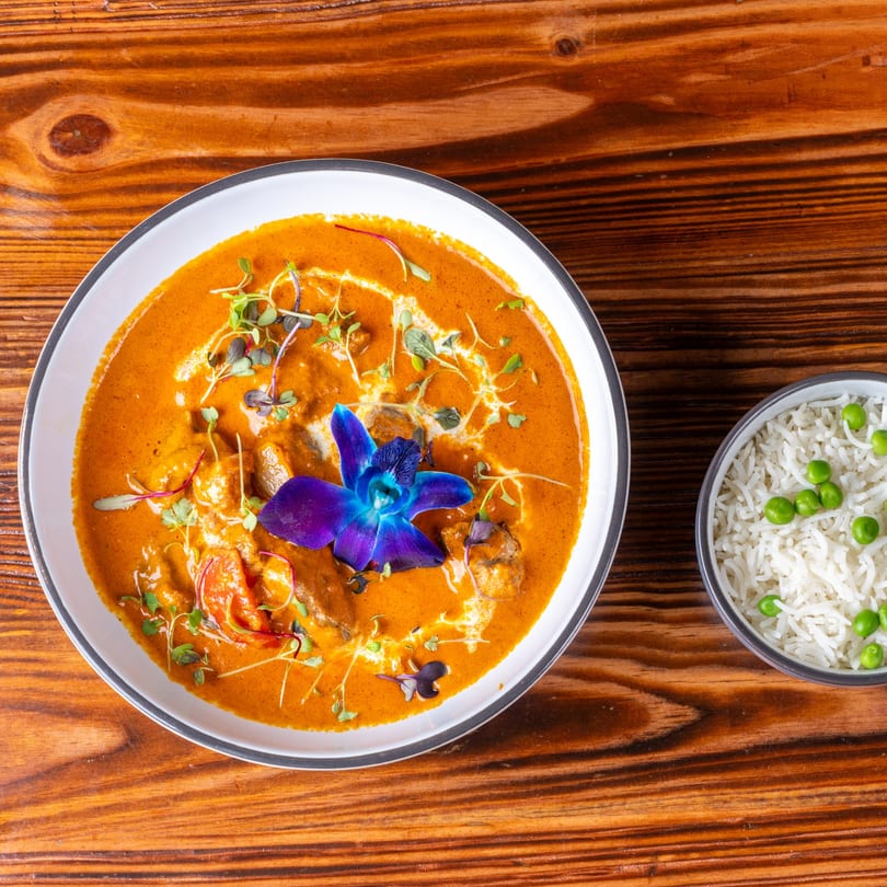 Orange curry with shrimp, microgreens, and a blue orchid, served with a side of rice and peas.